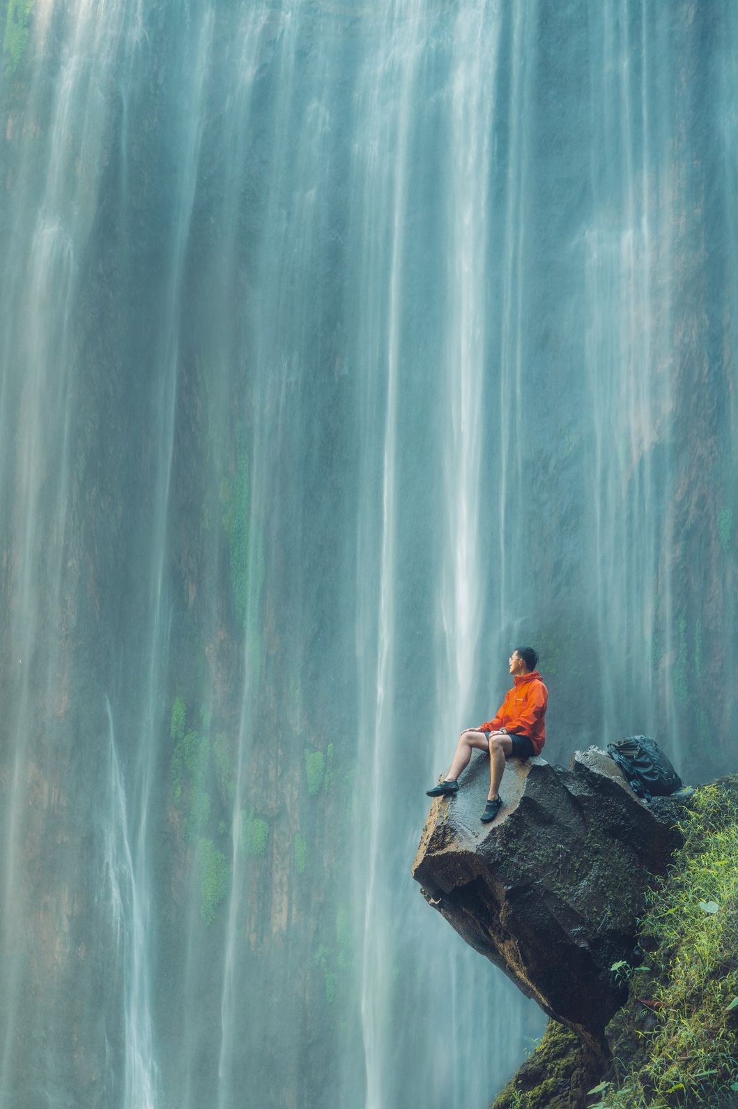 A person sitting on a rock near Tumpak Sewu Waterfall, surrounded by cascading water in East Java, Indonesia.