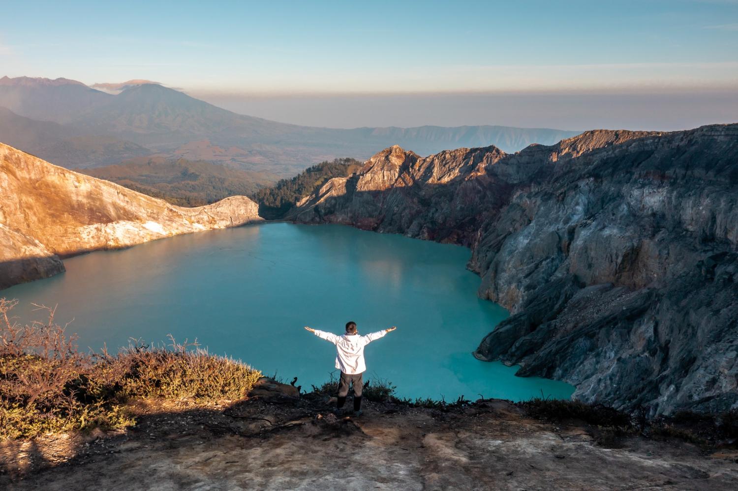 Tourist standing on the edge of Kawah Ijen crater, overlooking the turquoise lake and surrounding mountains.